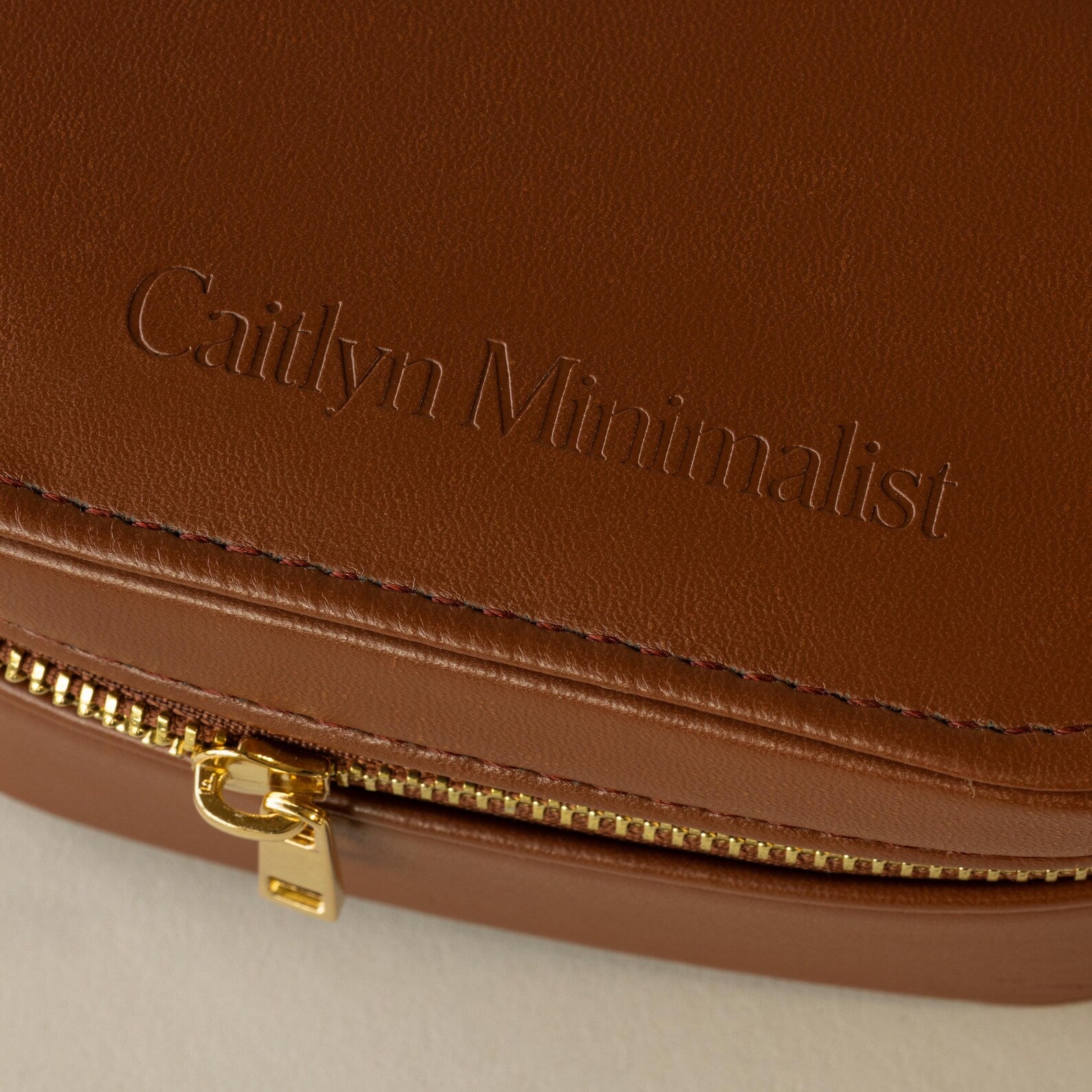 Close-up of an Oval Leather Jewelry Box, a brown leather pouch embossed with Caitlyn Minimalist and a gold zipper, perfect as a compact travel jewelry organizer.