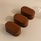 Three Oval Leather Jewelry Boxes in brown are lined up on a beige surface, each zippered case serving as a stylish and compact travel jewelry organizer.