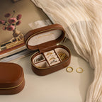 The Oval Leather Jewelry Box is a compact brown case perfect for storing gold rings, earrings, and bangles. Ideal for travel, it keeps your treasures organized and safe beside your book or other essentials.