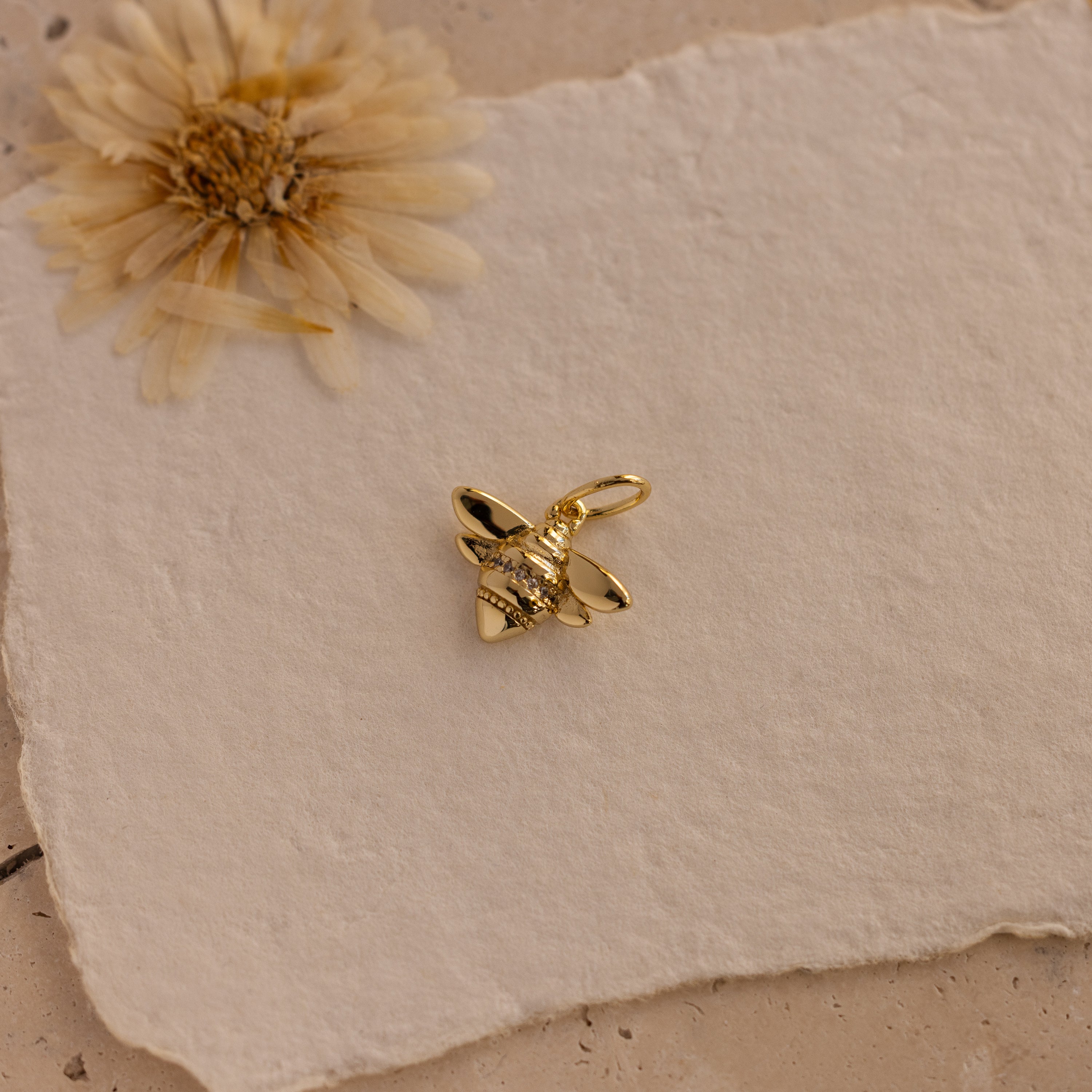 The Honeybee Charm in 18K Gold is displayed on textured white paper with a dried flower accent—an ideal gift for nature jewelry lovers.