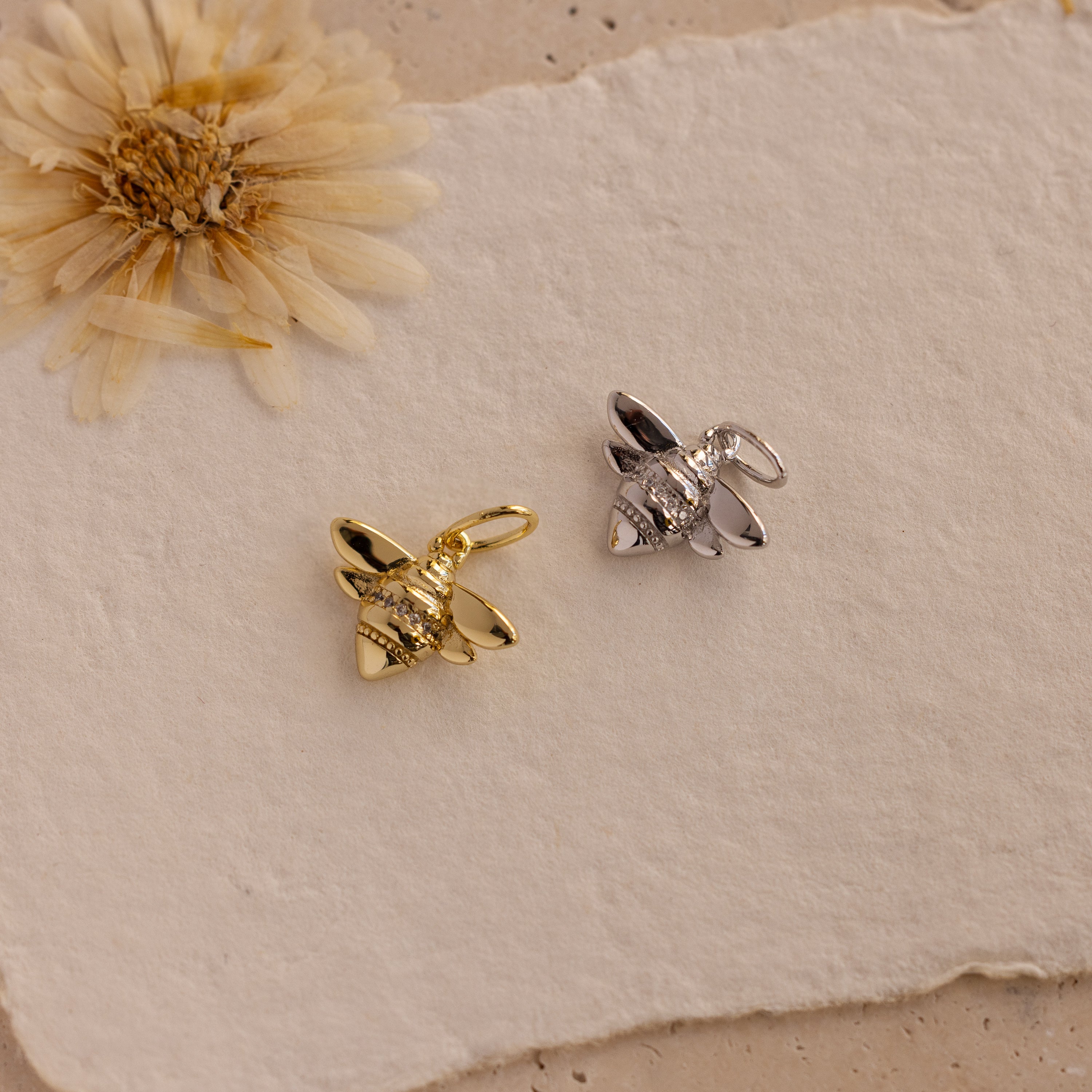 Honeybee Charm pendants, crafted in gold and silver bee shapes, are displayed on textured beige paper with a dried flower—an ideal nature-inspired accessory.