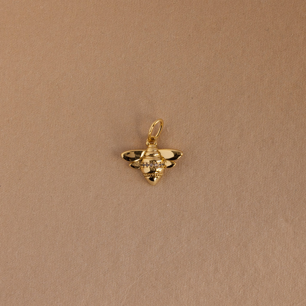 The Honeybee Charm in 18K Gold, adorned with small gemstones, is shown against a plain light brown background.