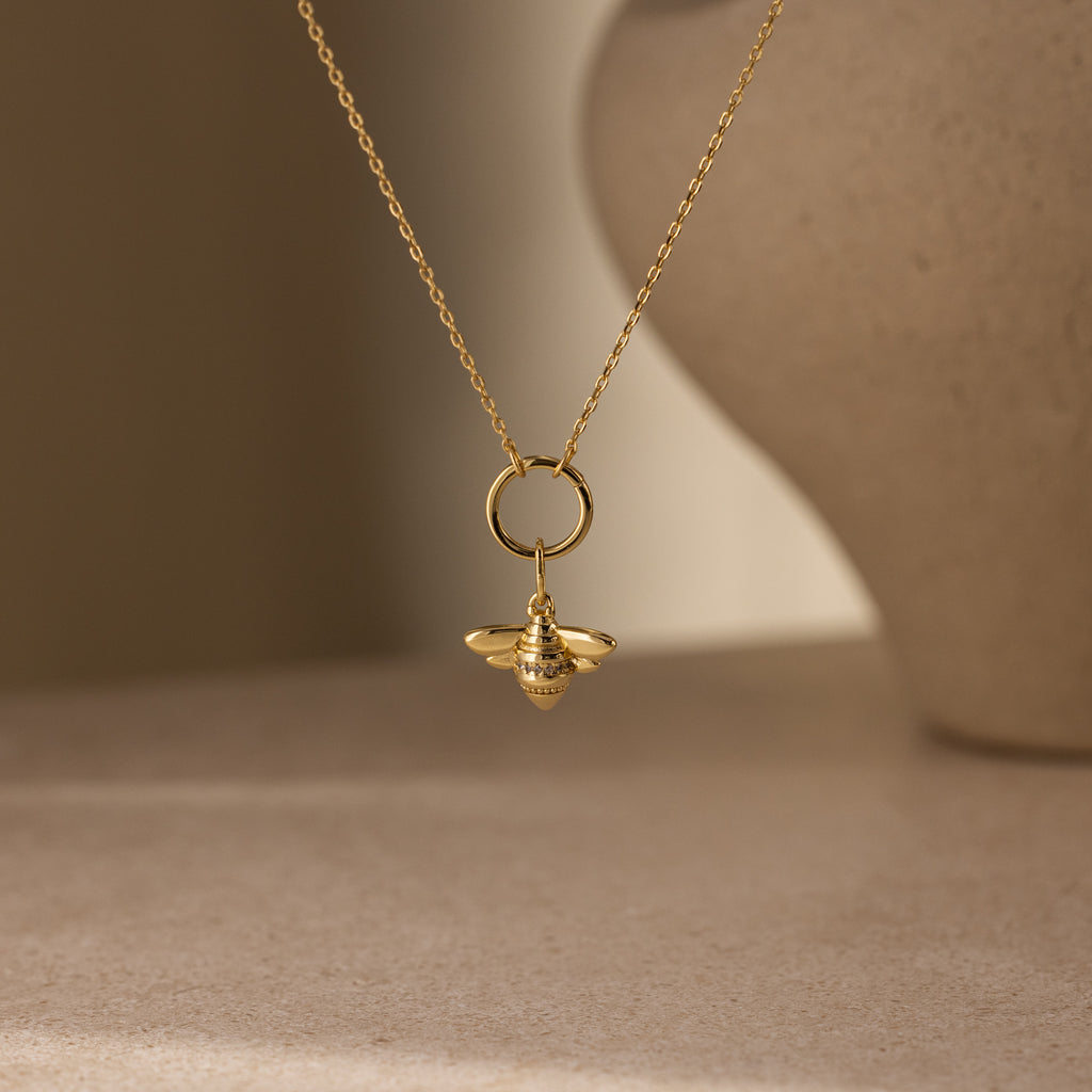 The Honeybee Charm in 18K Gold necklace features a delicate bee pendant, showcased against a softly blurred, neutral-toned background.