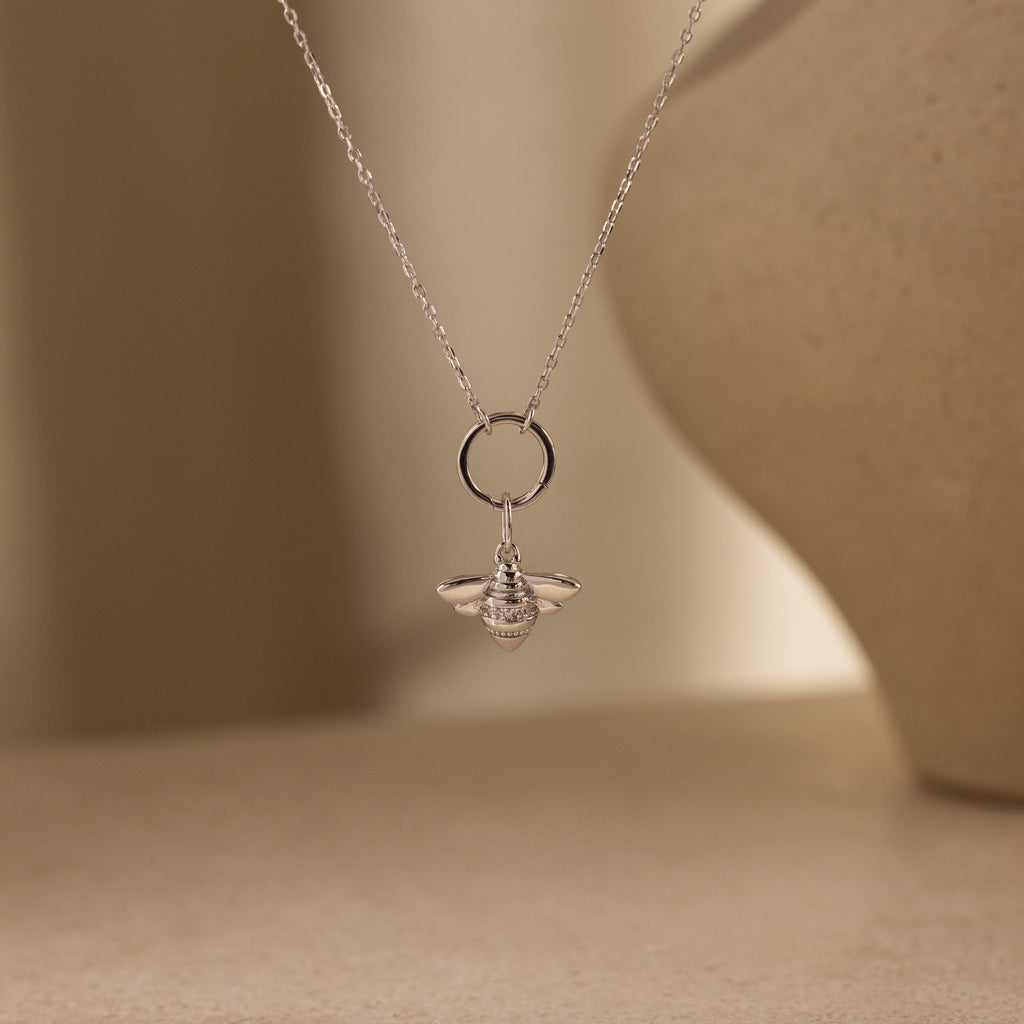 The Honeybee Charm in Sterling Silver hangs from a delicate chain necklace, highlighting exquisite nature-inspired jewelry against a neutral background.