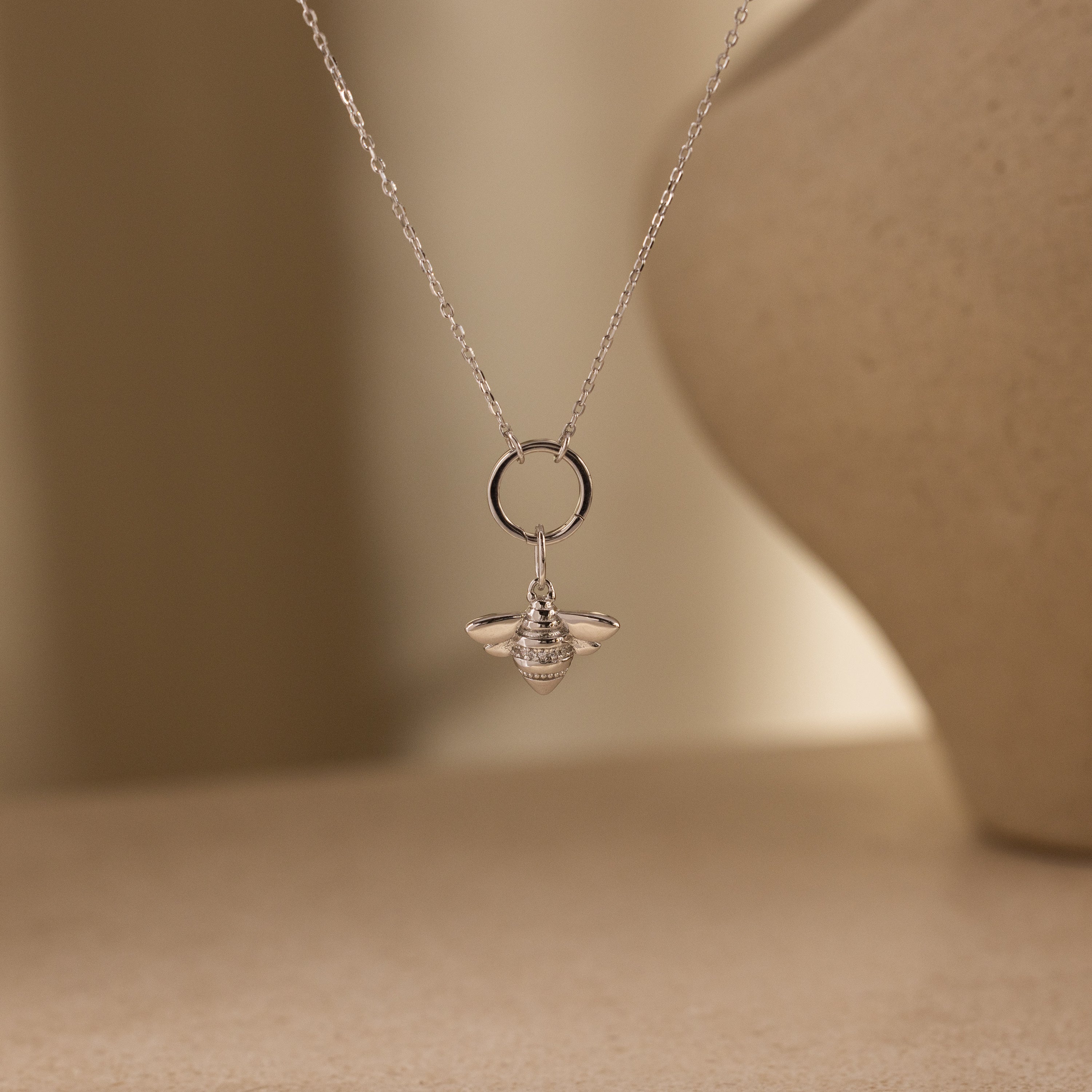The Honeybee Charm in Sterling Silver hangs from a delicate chain necklace, highlighting exquisite nature-inspired jewelry against a neutral background.