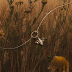 The Honeybee Charm is a silver pendant necklace on a chain, shown against wild grasses and yellow flowers—an elegant nature-inspired jewelry piece.
