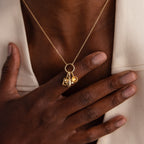 A hand touches a gold necklace featuring the Honeybee Charm pendant, worn over a cream-colored garment.