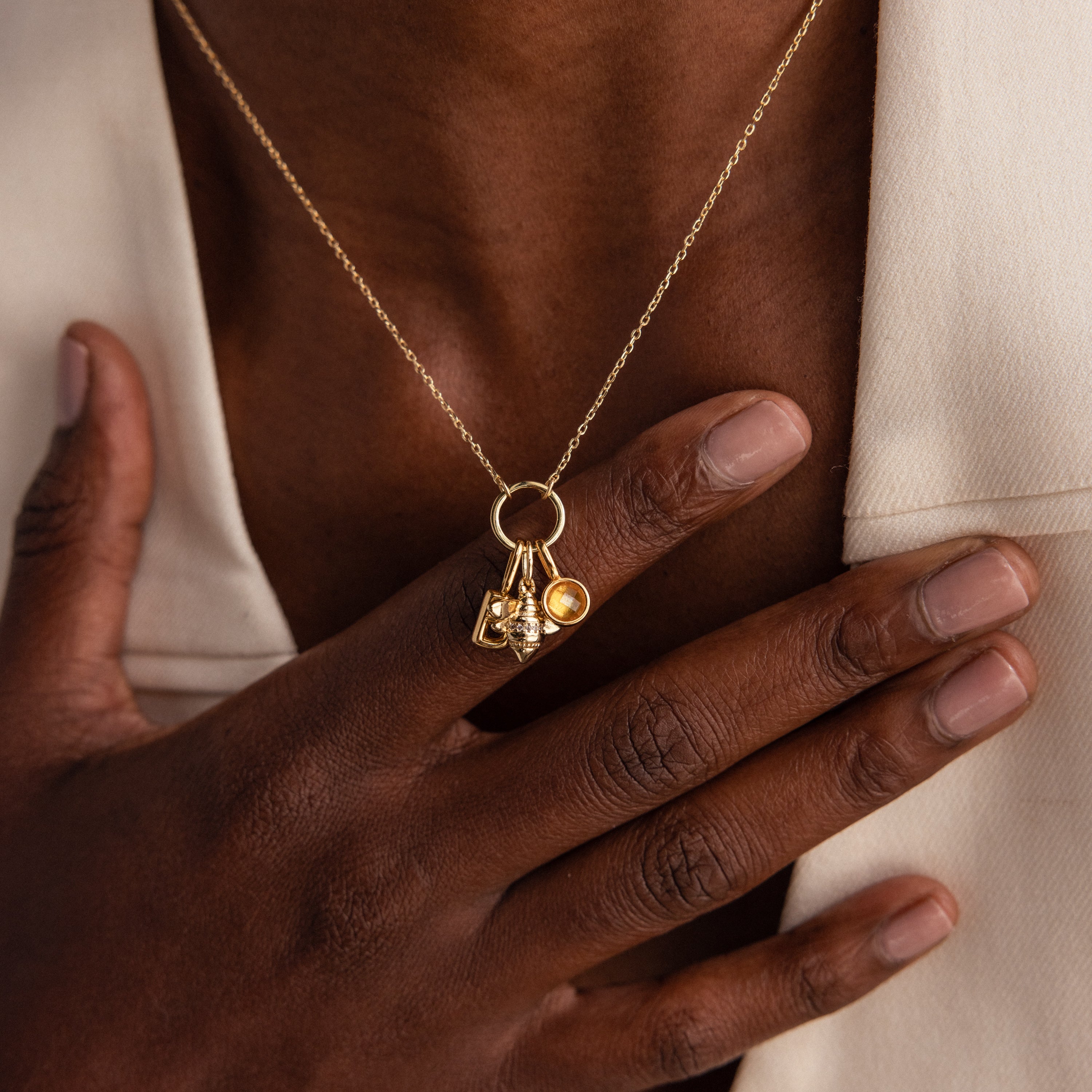 A hand touches a gold necklace featuring the Honeybee Charm pendant, worn over a cream-colored garment.