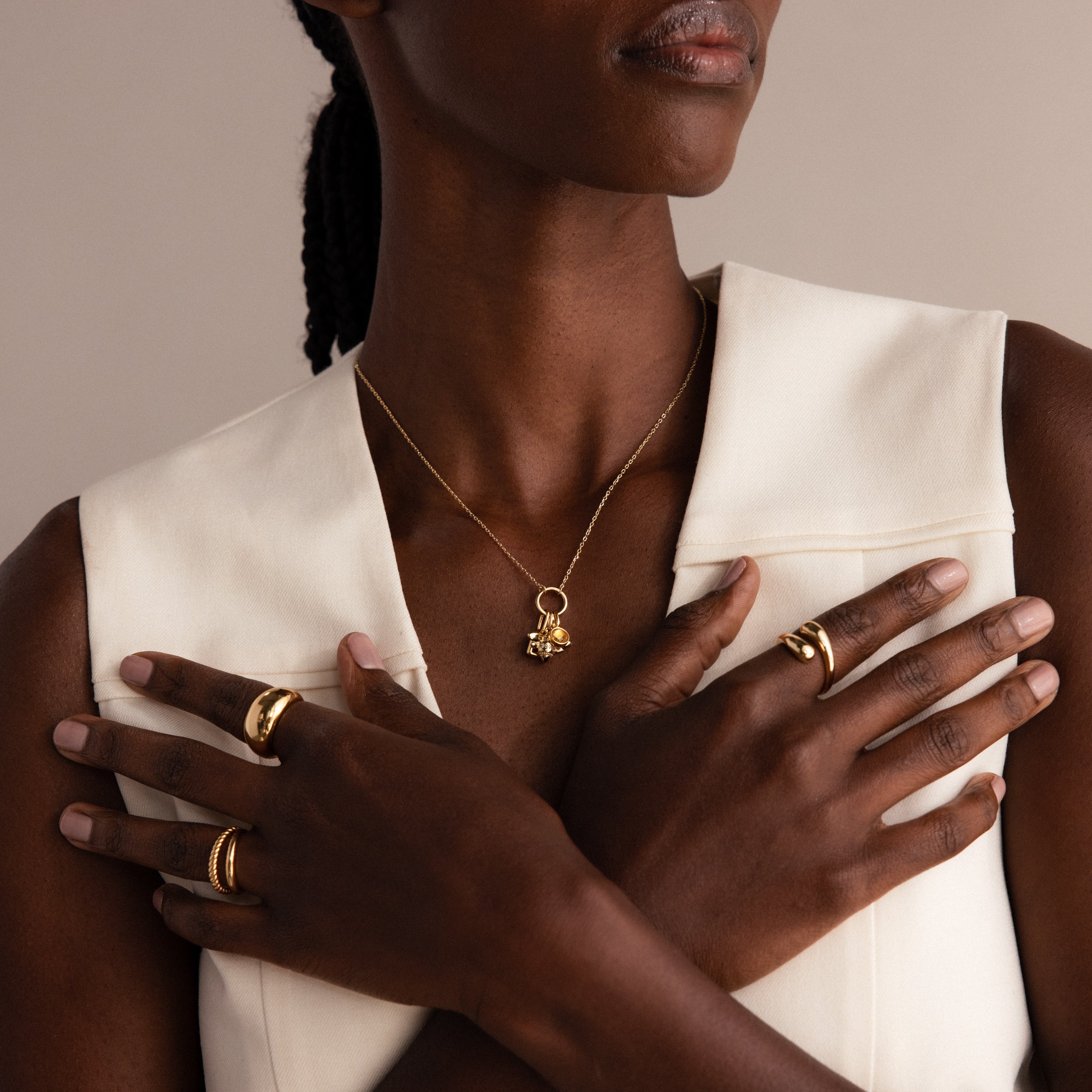 Woman in a sleeveless cream top wearing nature-inspired jewelry—a pavé band, gold rings, and the Honeybee Charm necklace—arms crossed in front of her chest.