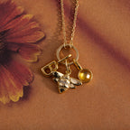 The Honeybee Charm in 18K Gold necklace features a letter B and yellow gem charms, shown on a floral-patterned background.