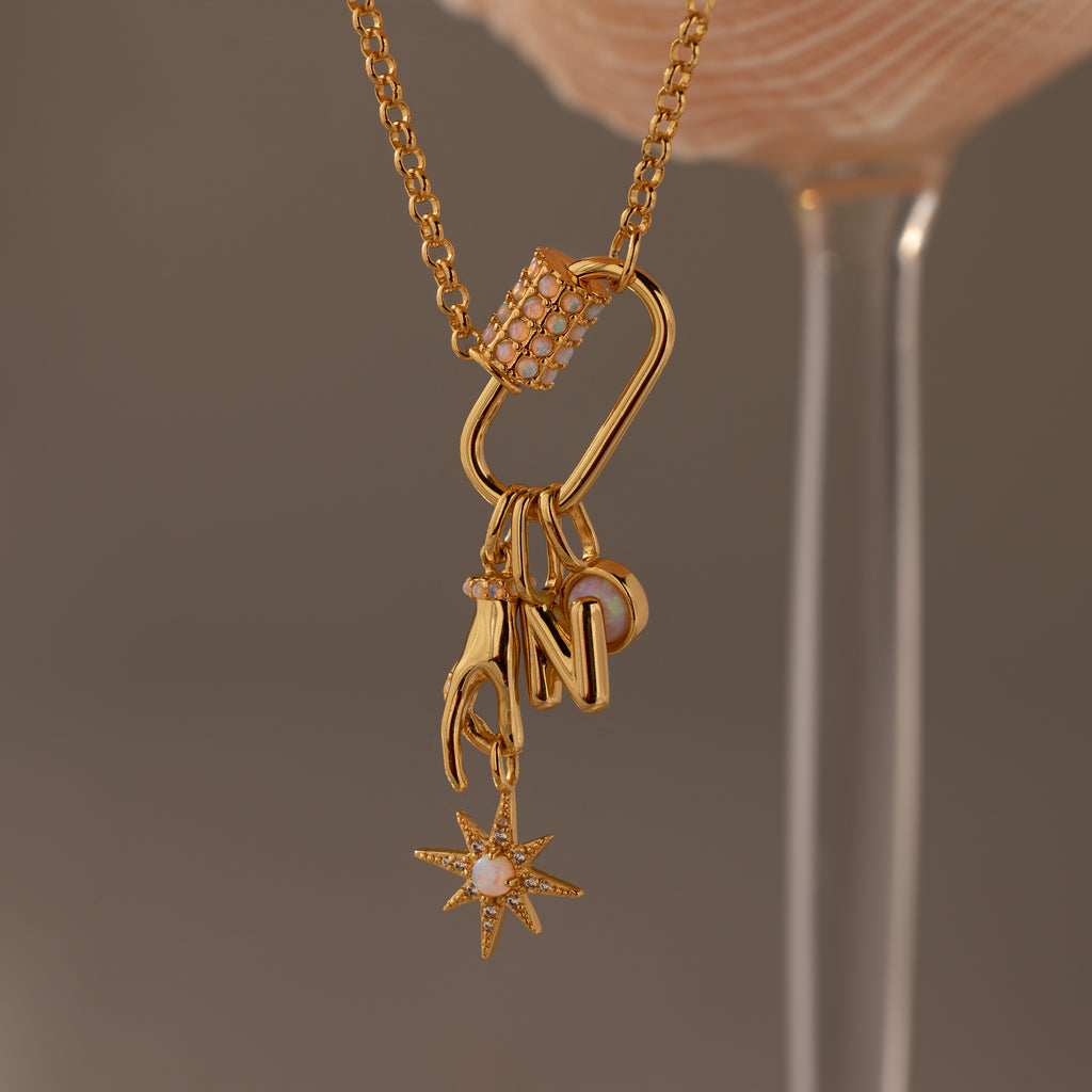 Gold necklace with charms—a hand, letter N, pink crystal bar, and starburst—hangs from a chain featuring the Opal Carabiner Clasp in 18K Gold, adding a modern twist to this playful charm connector style.