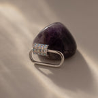 A white gold, oval-shaped Opal Carabiner Clasp with gems is displayed on a polished amethyst stone atop a beige surface.