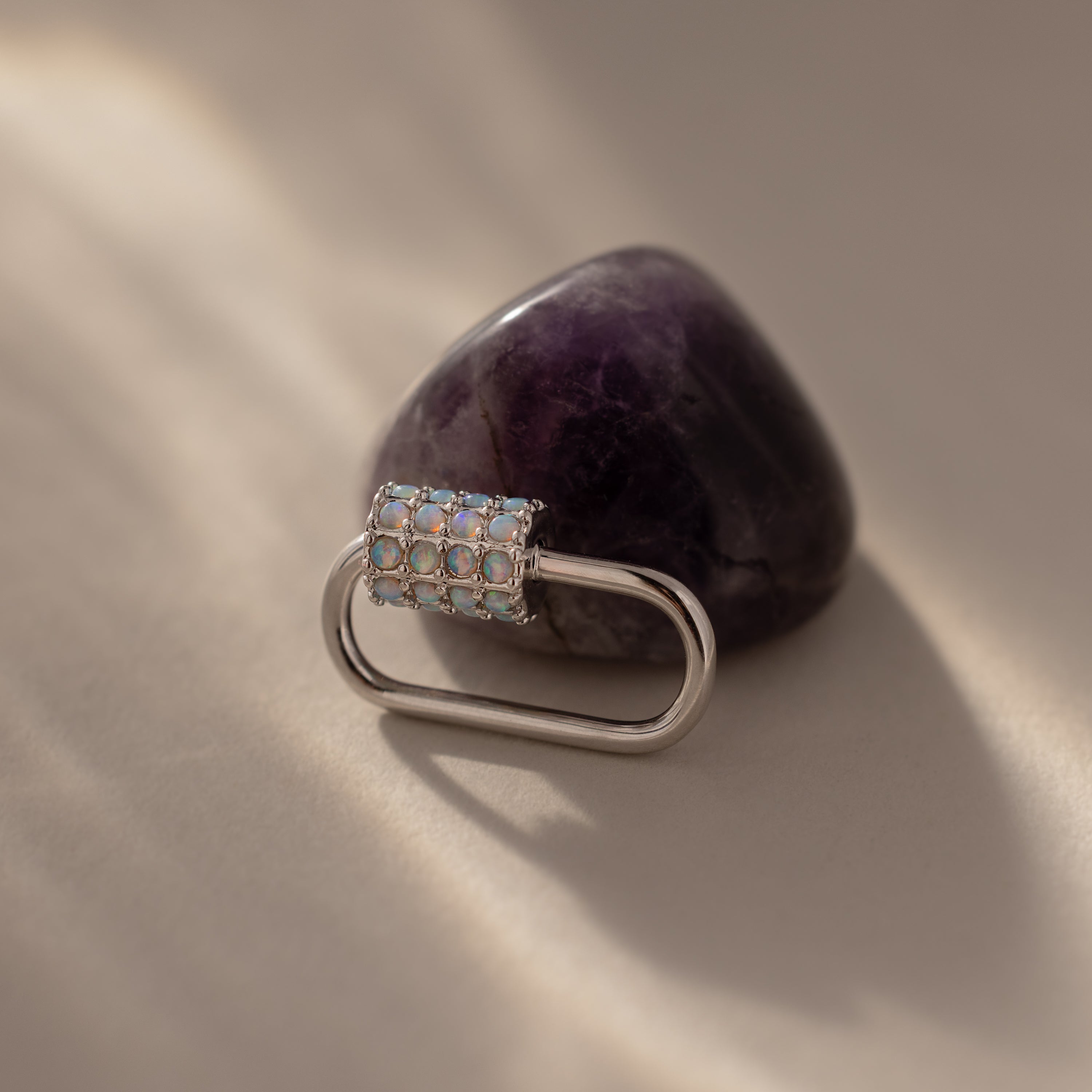 The Opal Carabiner Clasp, adorned with opal-like stones, is displayed against a polished purple amethyst stone on beige.