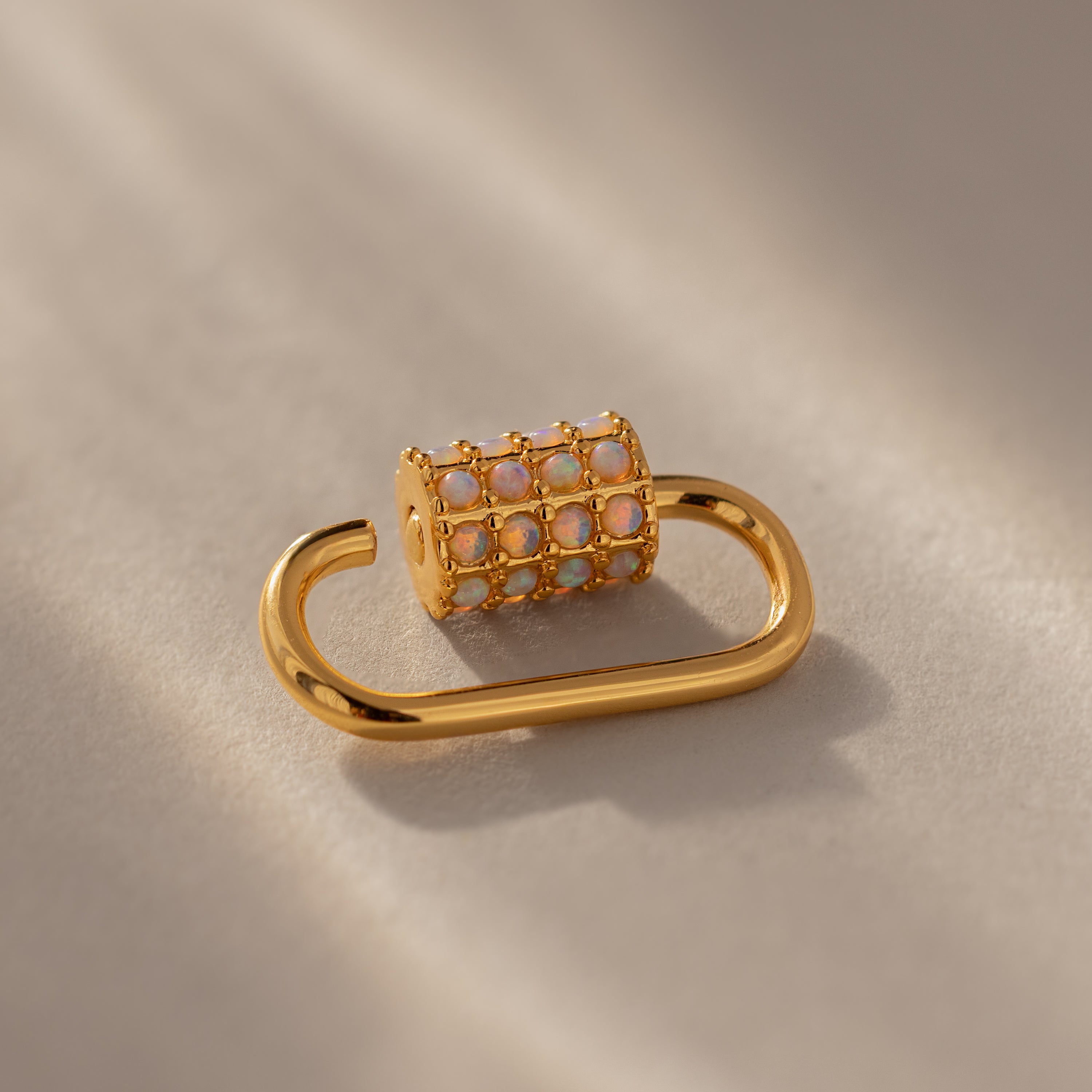 The Opal Carabiner Clasp is a gold earring featuring a cylindrical charm connector set with small opal-like stones, displayed on a neutral background.