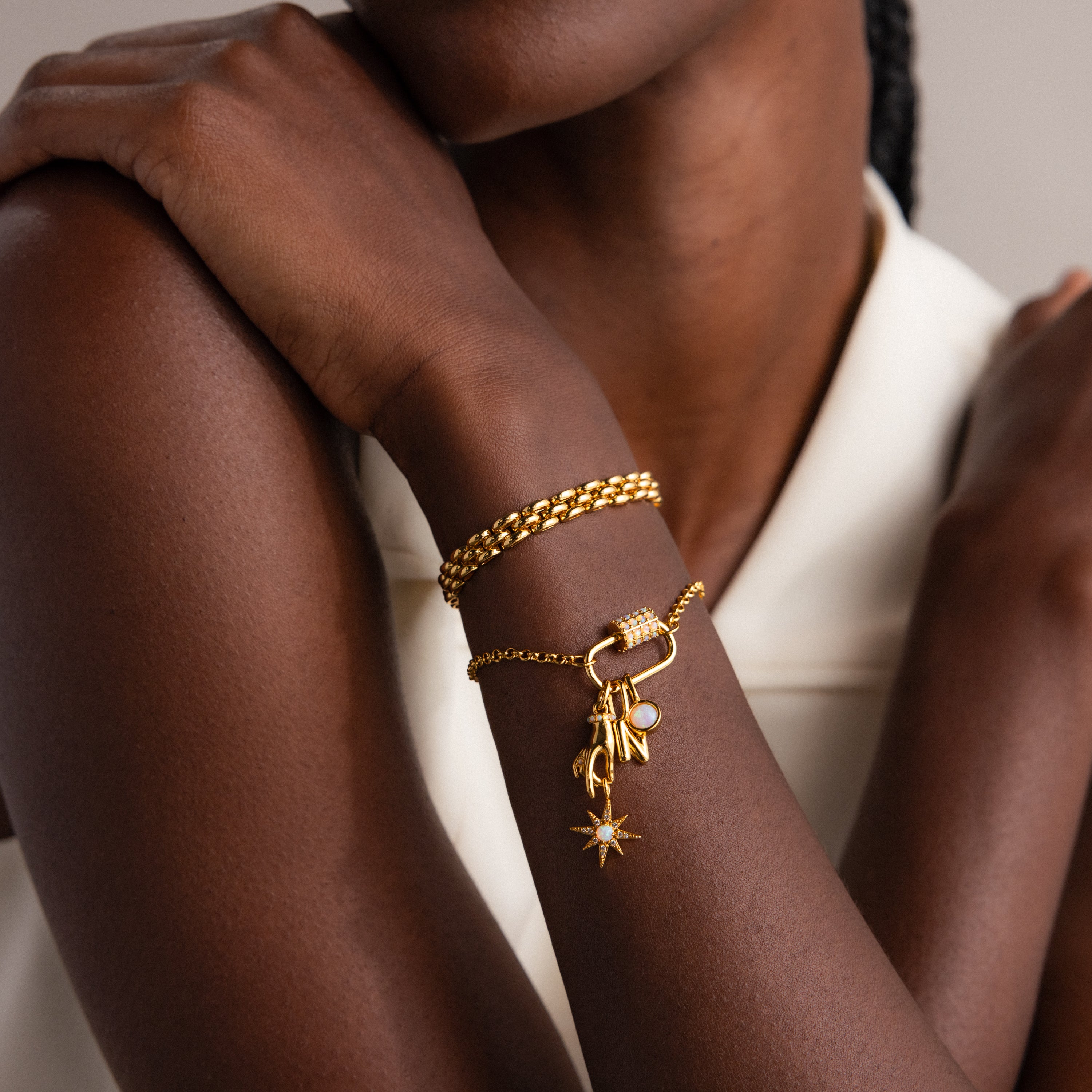 A woman in a white sleeveless top crosses her arms, wearing gold bracelets including one featuring the Opal Carabiner Clasp in 18K Gold.