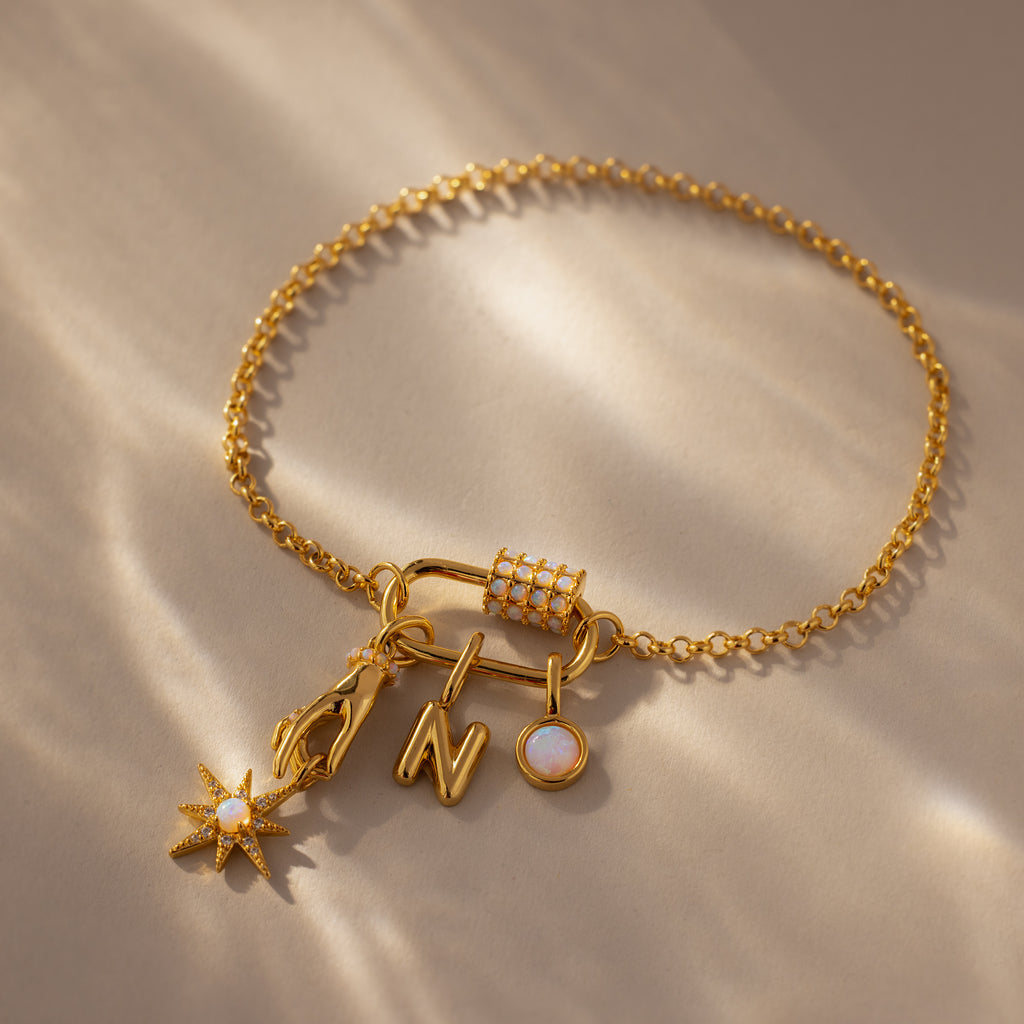 The Opal Carabiner Clasp in 18K Gold secures a gold chain bracelet with four charms—a star, lightning bolt, letter N, and round gem—offering a unique modern style for opal jewelry lovers. Shown on a stylish beige surface.