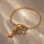 The Opal Carabiner Clasp in 18K Gold secures a gold chain bracelet with four charms—a star, lightning bolt, letter N, and round gem—offering a unique modern style for opal jewelry lovers. Shown on a stylish beige surface.