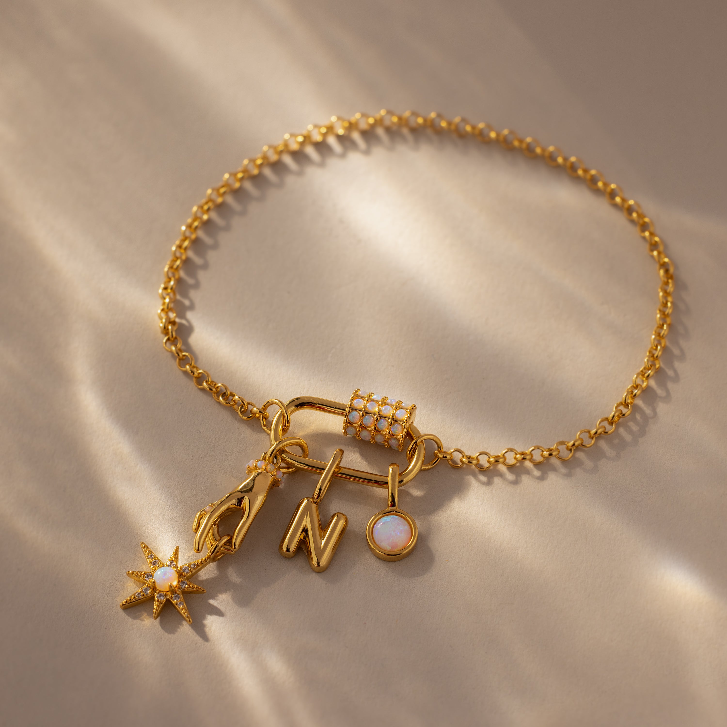 The Opal Carabiner Clasp in 18K Gold secures a gold chain bracelet with four charms—a star, lightning bolt, letter N, and round gem—offering a unique modern style for opal jewelry lovers. Shown on a stylish beige surface.