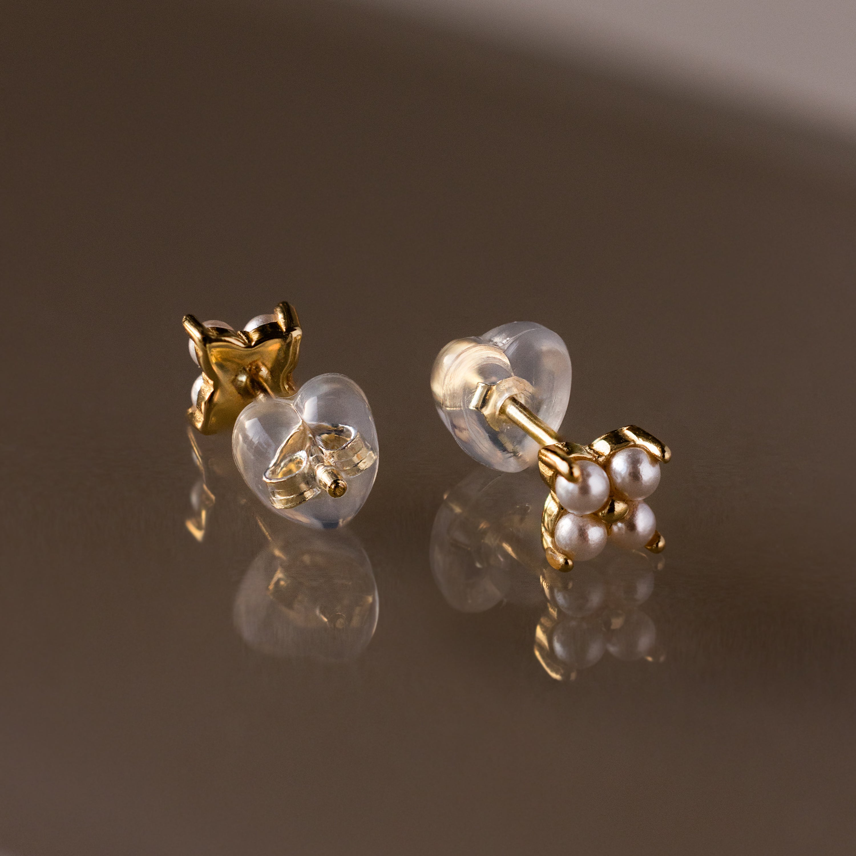 Heart & Flower Earring Backs secure your gold stud earrings with small white pearls—your earring essentials—shown on a reflective brown surface.