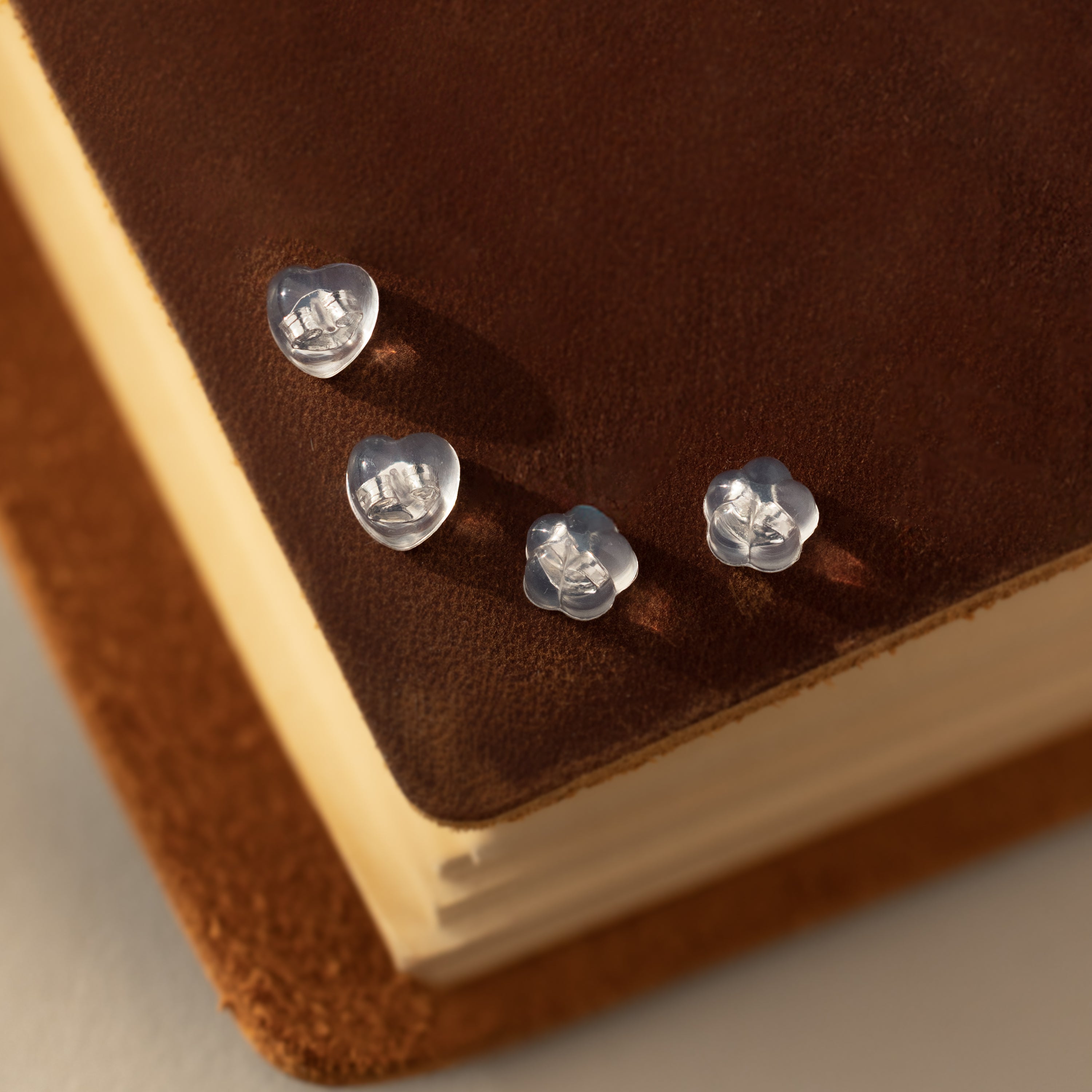 Four Heart & Flower Earring Backs in Sterling Silver, must-have essentials for any jewelry collection, are displayed on a brown suede-covered book.