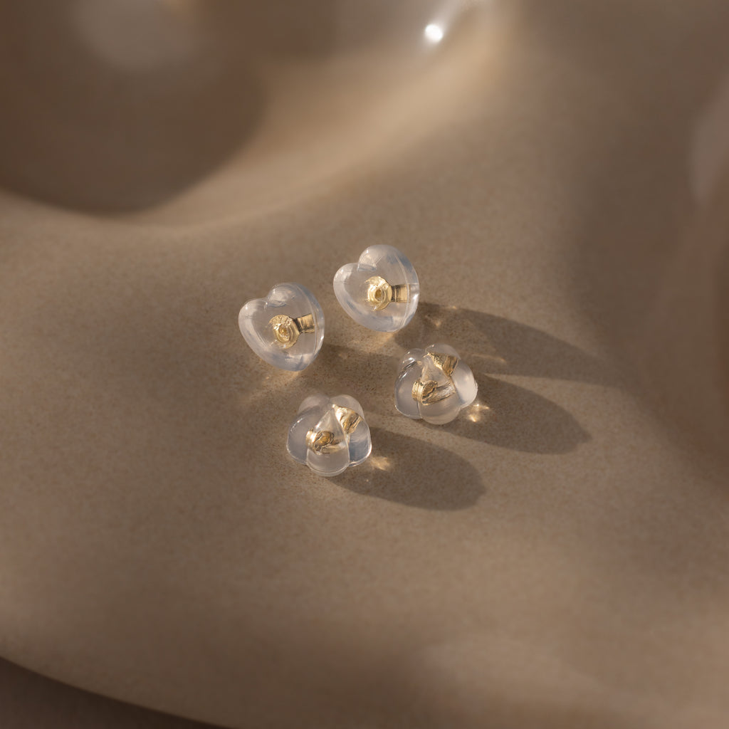Four clear heart-shaped earrings with 18K gold studs and silicone backs—Heart & Flower Earring Backs in 18K Gold—are displayed on a smooth, beige surface.
