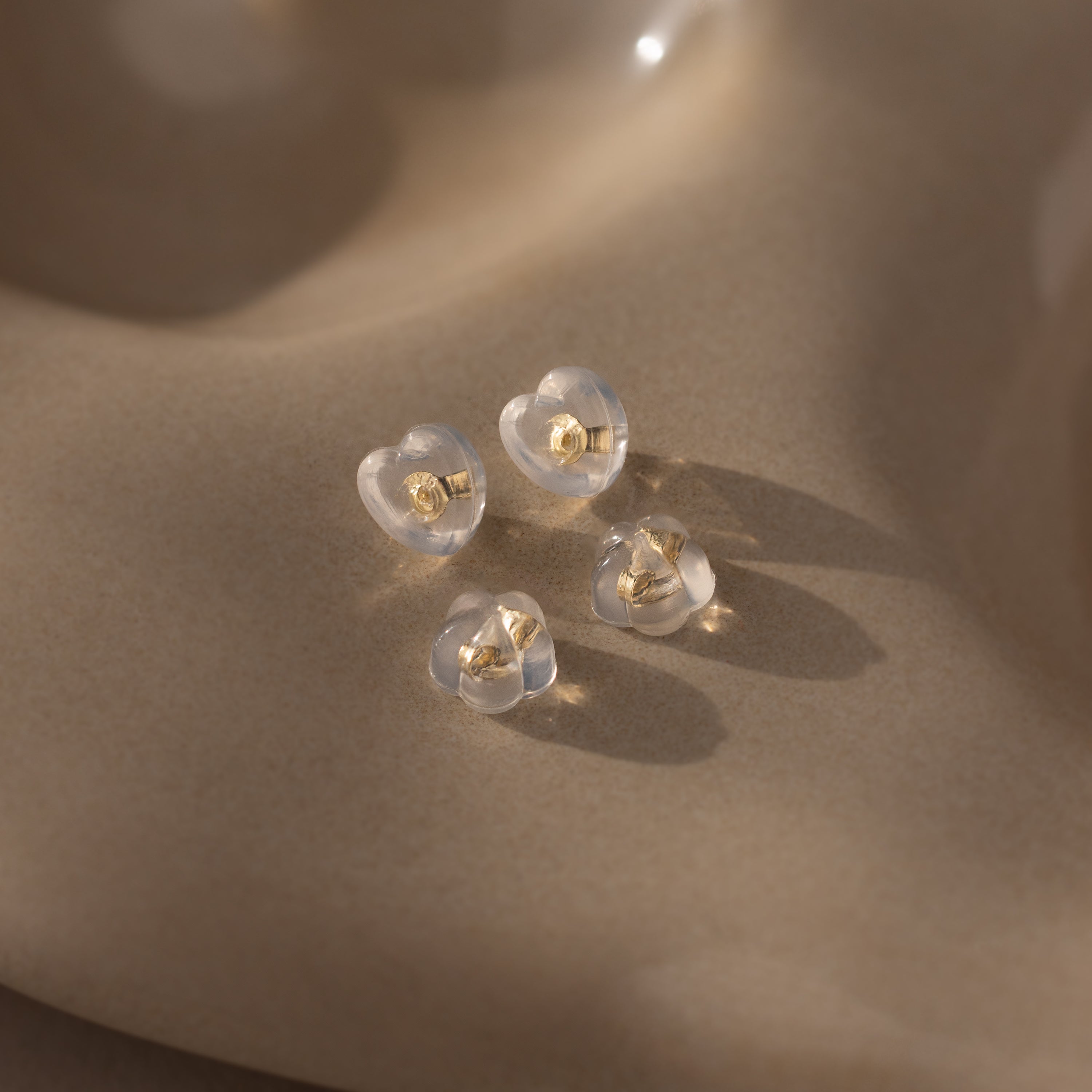 Four clear heart-shaped earrings with 18K gold studs and silicone backs—Heart & Flower Earring Backs in 18K Gold—are displayed on a smooth, beige surface.