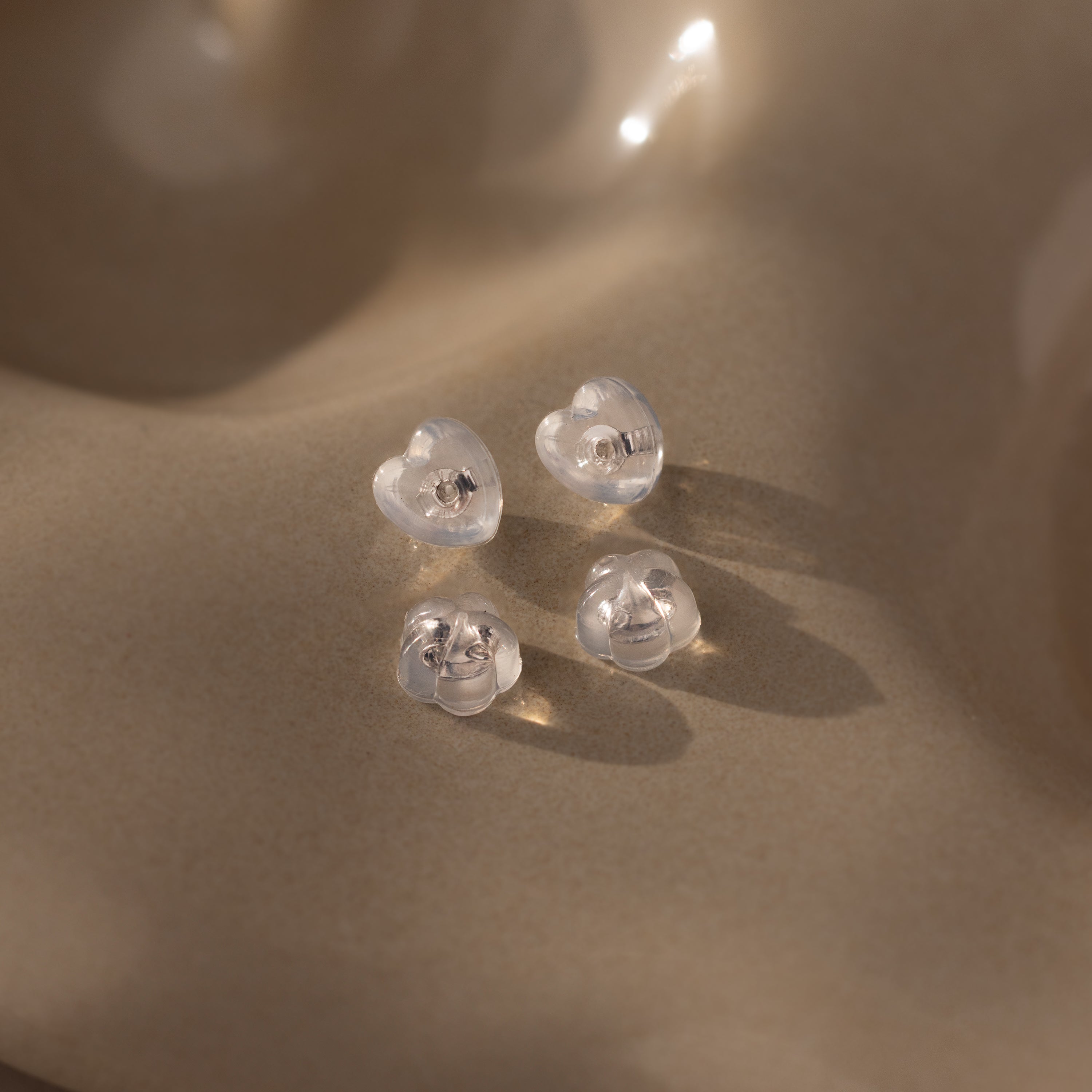 Heart & Flower Earring Backs in Sterling Silver—featuring clear heart and round silicone studs—rest on a smooth beige surface in soft lighting, ensuring secure and comfortable wear.