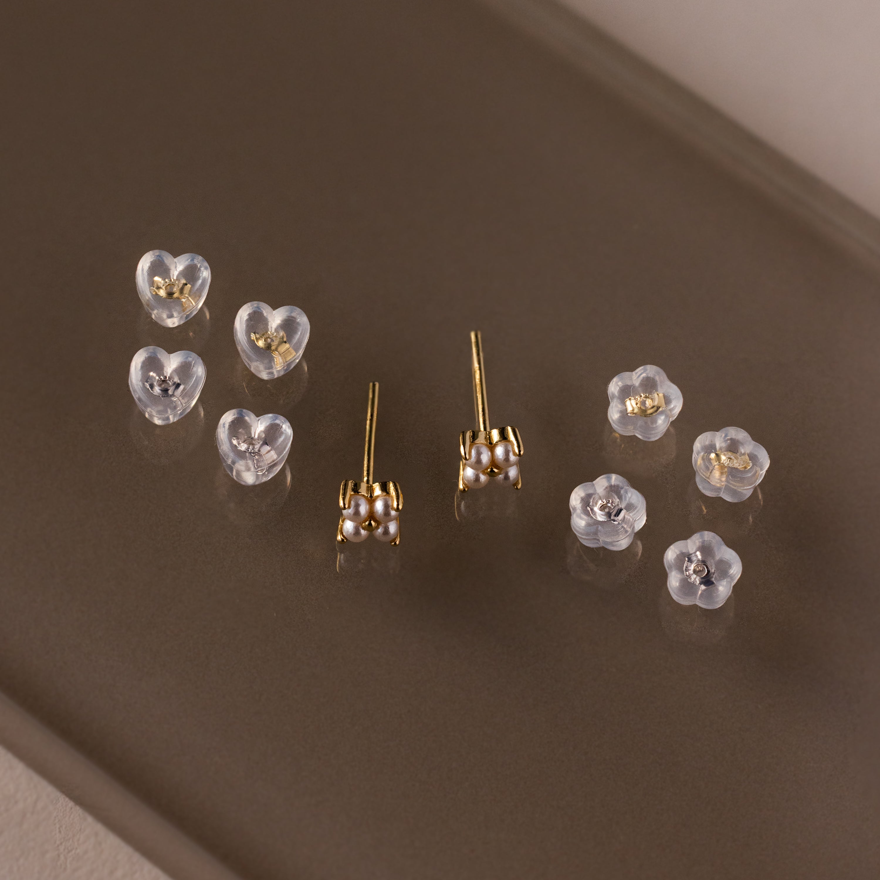 Five pairs of delicate Heart & Flower Earring Backs in 18K Gold, each with secure silicone stud backs and elegant gold and pearl details—perfect essentials, displayed on a brown surface.