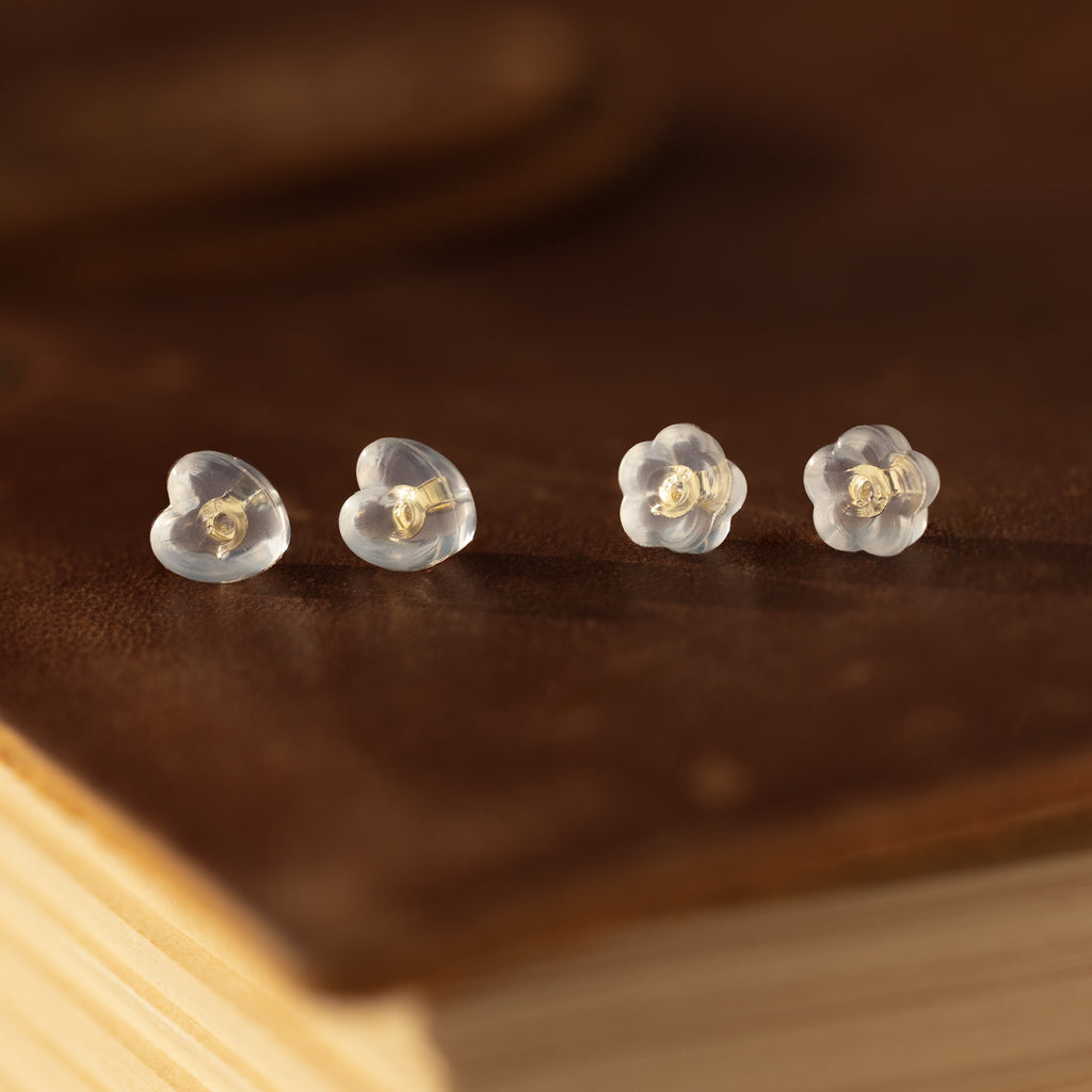 Two pairs of earring essentials: Heart & Flower Earring Backs in 18K Gold, displayed on a brown surface.
