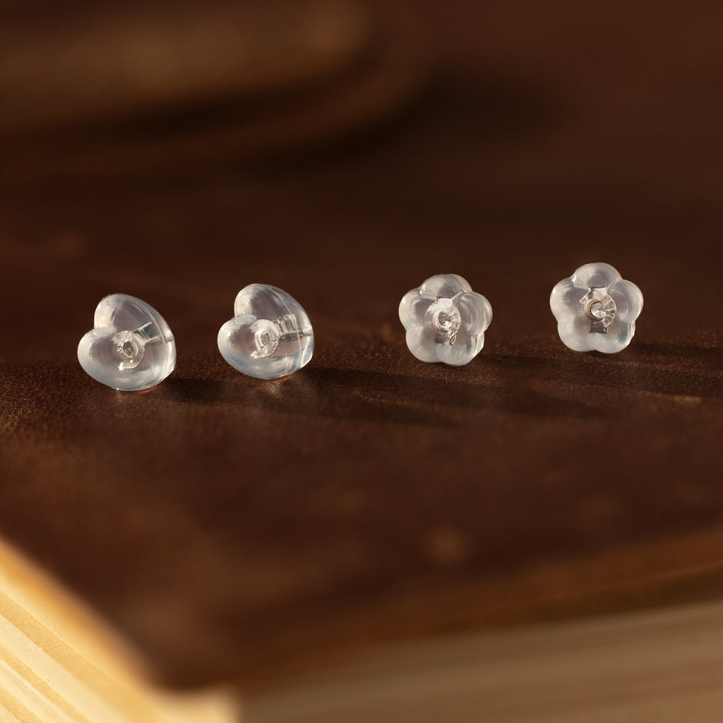 Two pairs of clear heart and flower-shaped earrings with sterling silver backs—Heart & Flower Earring Backs in Sterling Silver—displayed on a brown leather surface; perfect earring essentials.