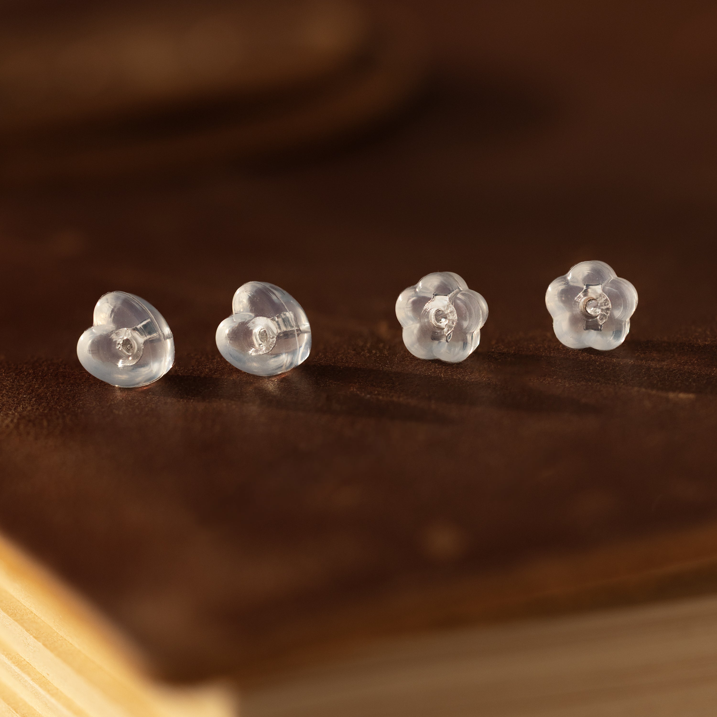 Two pairs of clear heart and flower-shaped earrings with sterling silver backs—Heart & Flower Earring Backs in Sterling Silver—displayed on a brown leather surface; perfect earring essentials.