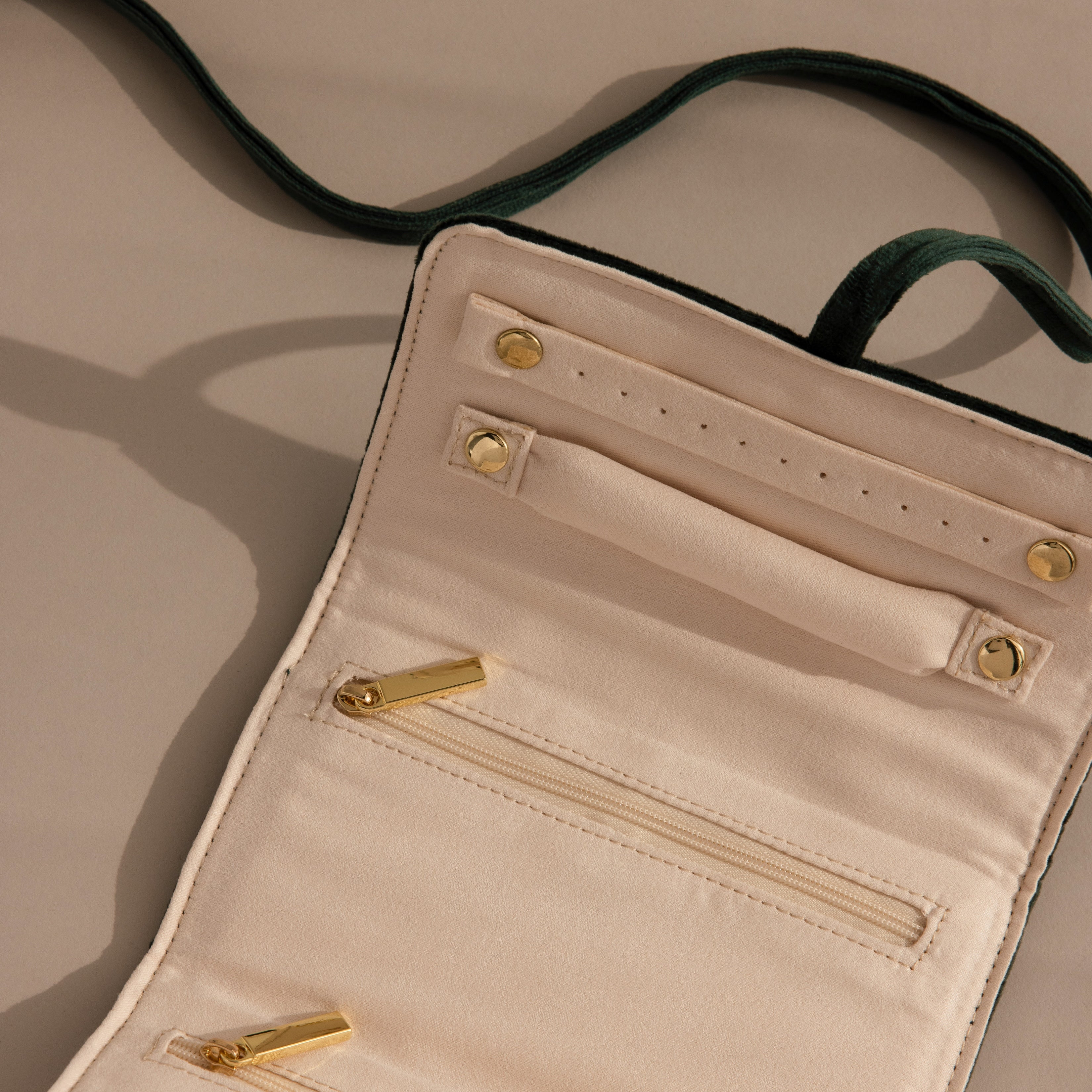 Open cream-colored Velvet Roll-Up Jewelry Case with gold zippers and buttons, lying on a beige surface with a visible green strap—an ideal stylish travel organizer for your jewelry essentials.