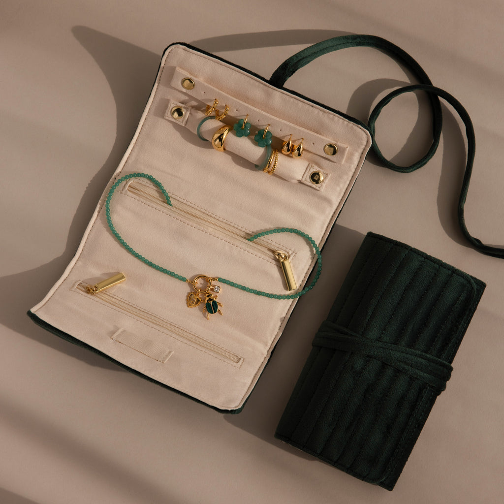 A Velvet Roll-Up Jewelry Case in green velvet is open to reveal gold and green jewelry, with its closed form beside it on a beige surface.
