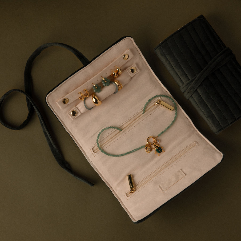 A Velvet Roll-Up Jewelry Case opens to display gold rings, earrings, and a green beaded necklace arranged on a dark surface.