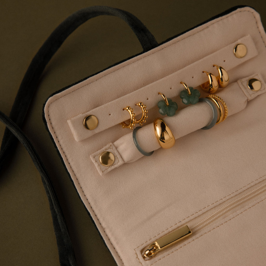 The Velvet Roll-Up Jewelry Case displays gold rings with green clover designs on a beige background and features a zipper pocket—ideal for organizing jewelry during travel.