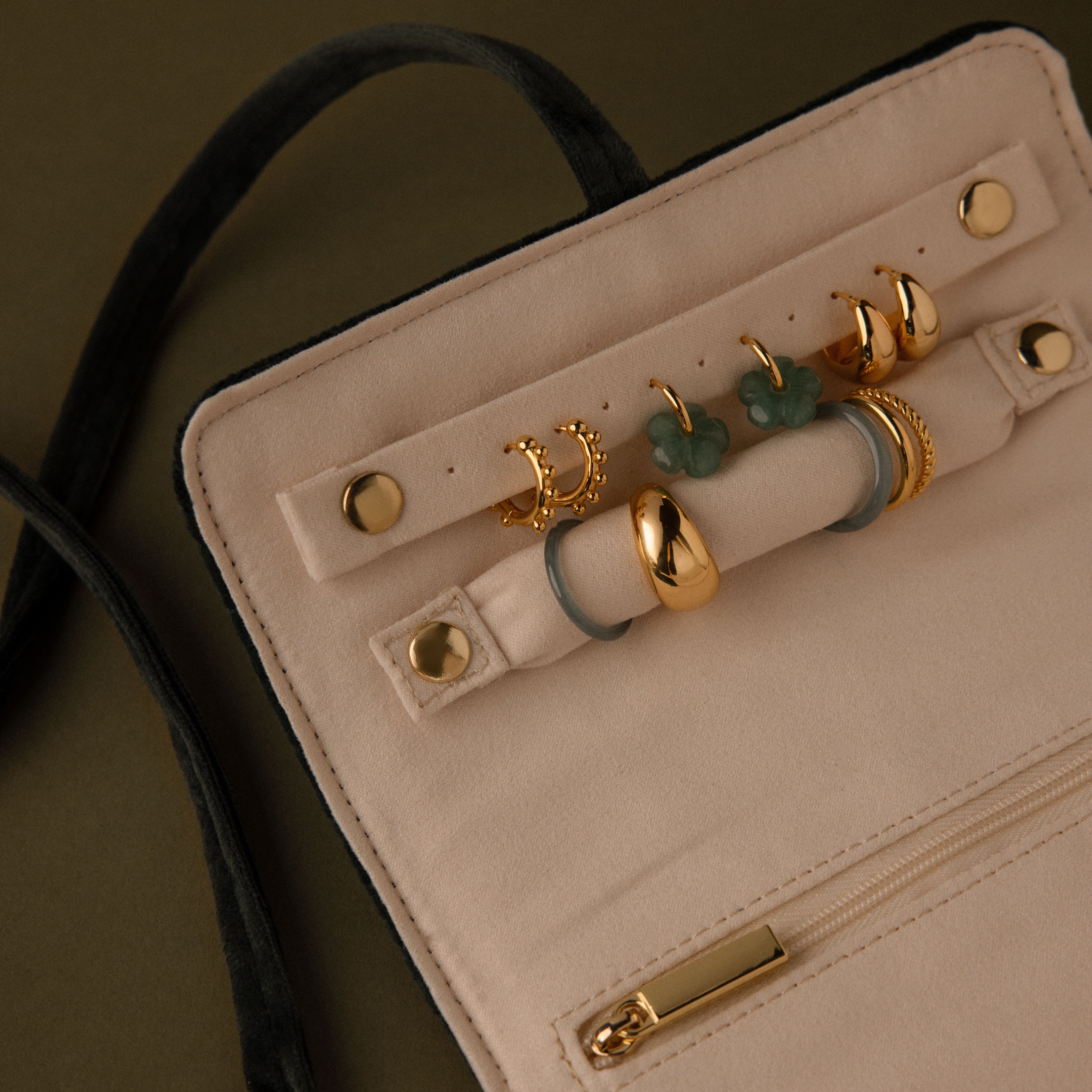 The Velvet Roll-Up Jewelry Case displays gold rings with green clover designs on a beige background and features a zipper pocket—ideal for organizing jewelry during travel.