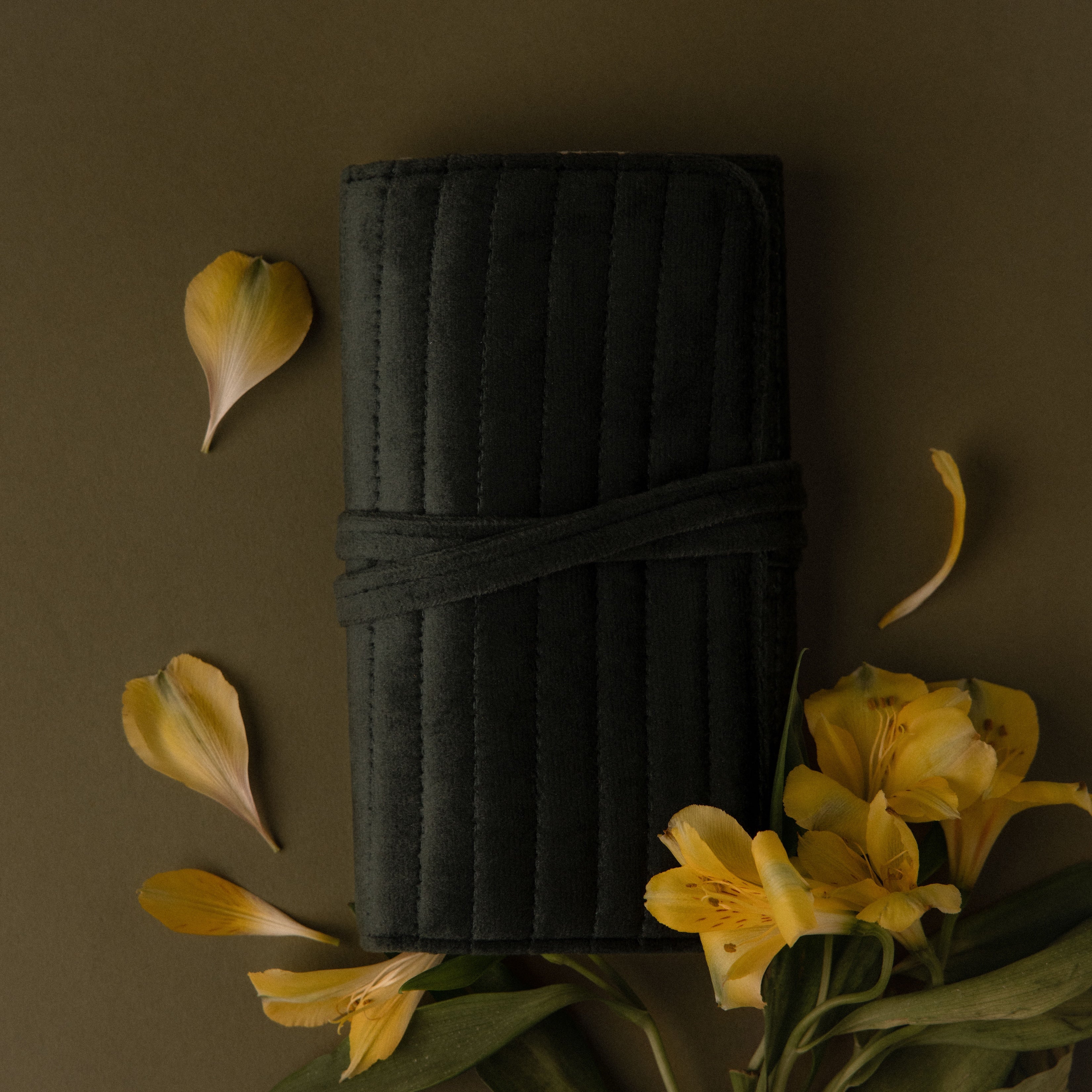 A Velvet Roll-Up Jewelry Case, surrounded by yellow flowers and petals, is displayed on an olive green background.