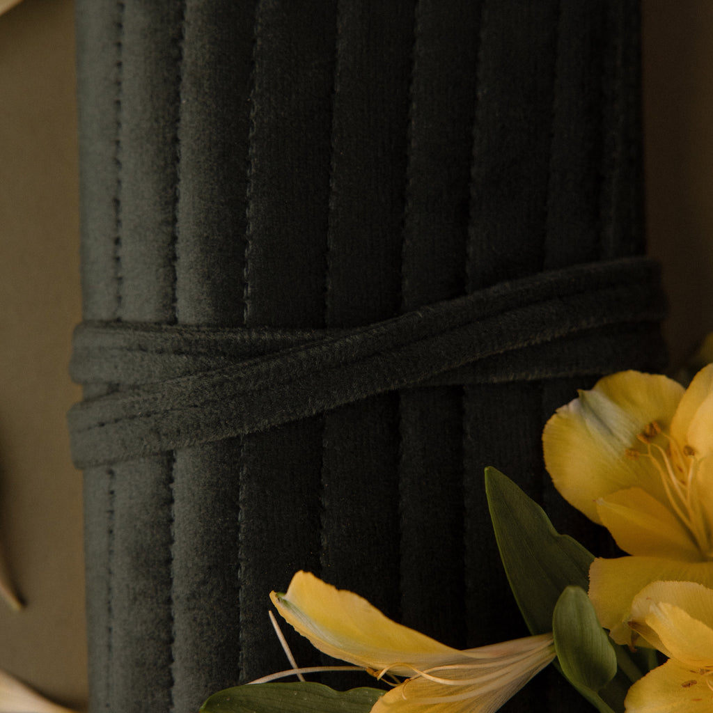 The Velvet Roll-Up Jewelry Case, made of dark green quilted fabric with a tie closure, is shown next to yellow flowers and green leaves.