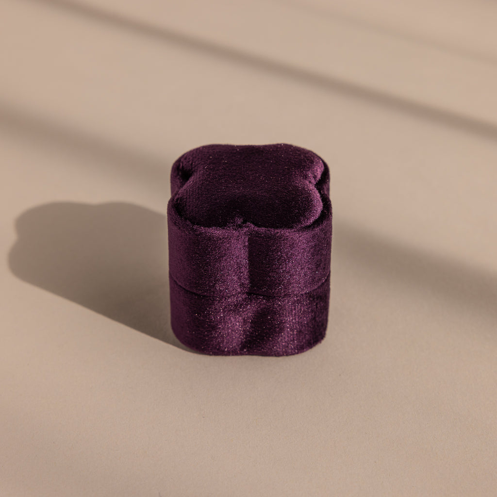 The Purple Flower Velvet Ring Box, a small purple box with a scalloped lid, sits on a beige surface with soft shadows.