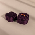 The Purple Flower Velvet Ring Box, ideal for proposals, is shaped like a flower and holds a sparkling ring inside as it sits on a beige surface.