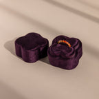 The Purple Flower Velvet Ring Box, ideal for proposals, is shaped like a flower and holds a sparkling ring inside as it sits on a beige surface.