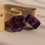 The Purple Flower Velvet Ring Box and band rest on a neutral background, accented by a wavy beige decorative element.