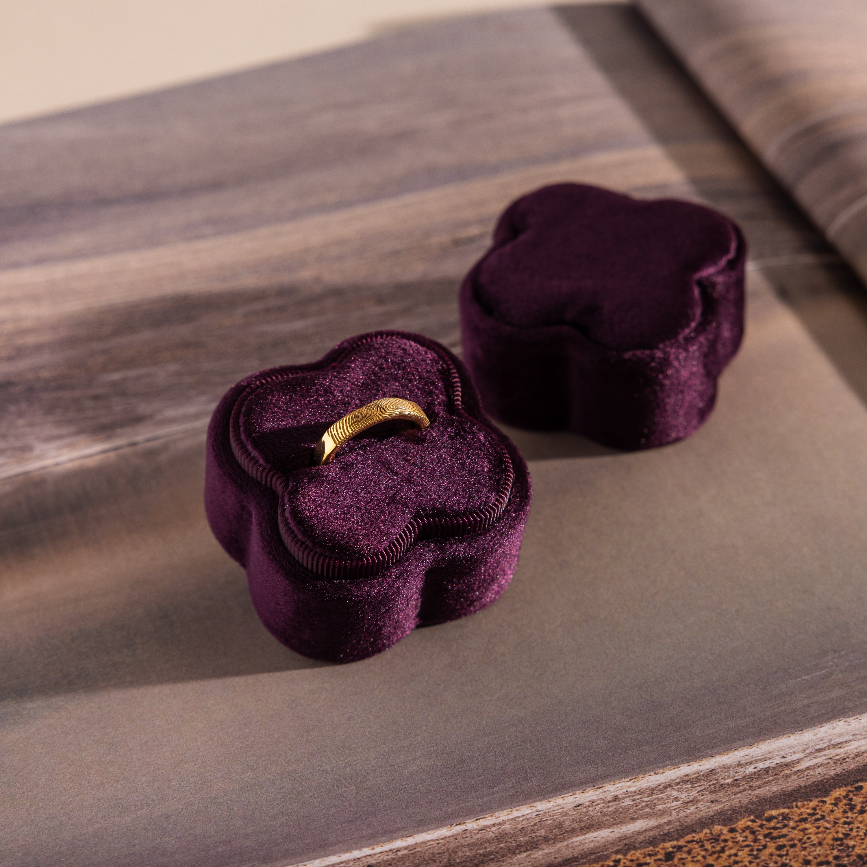 A gold ring rests in a Purple Flower Velvet Ring Box with its lid beside it on a textured surface, the box's deep purple color enhancing the elegant, dreamy scene.