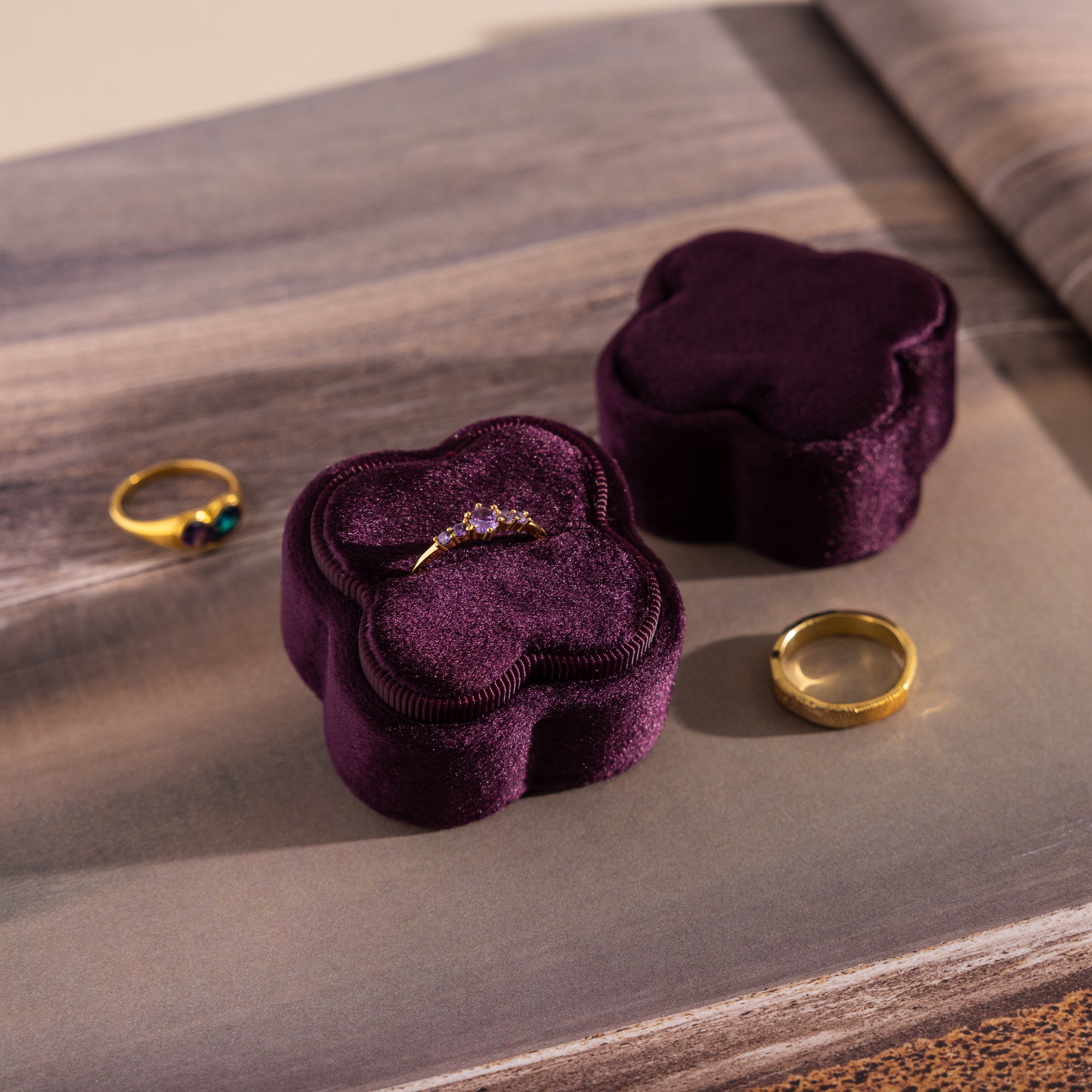 The Purple Flower Velvet Ring Box, shown with a gold band and a green stone ring beside it, offers an elegant way to present your proposal ring for that unforgettable special moment.