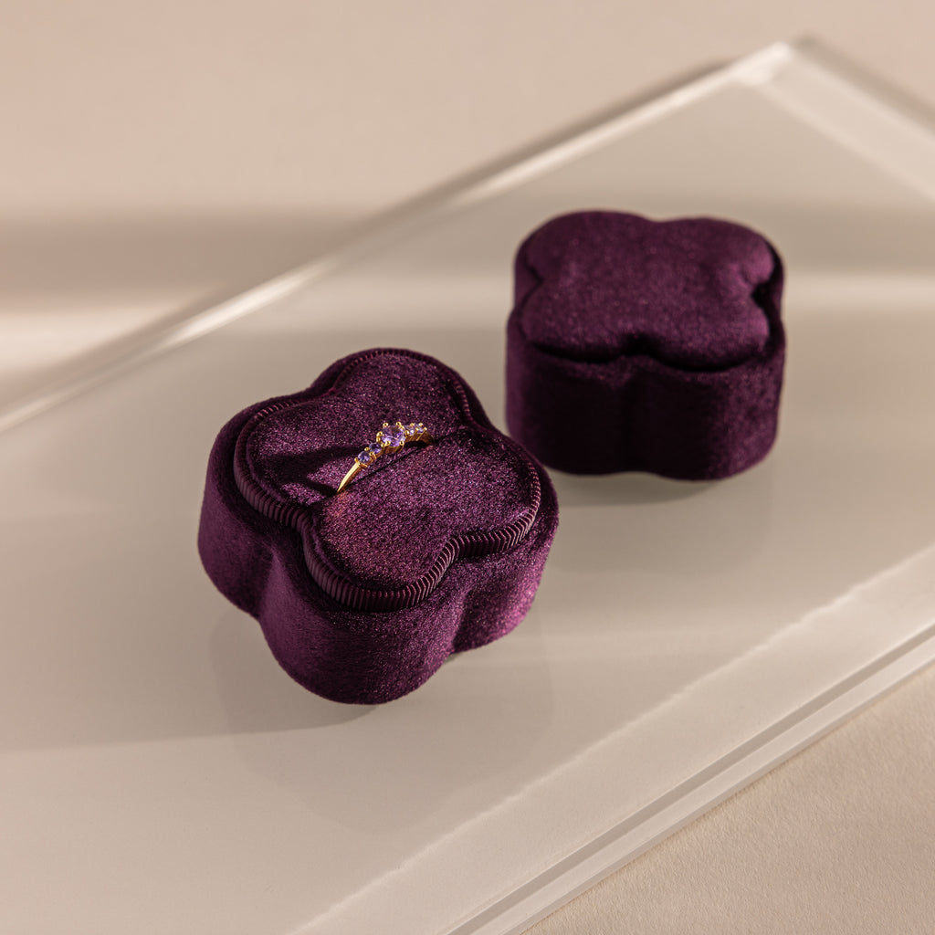 A gold ring with purple stones sits in the Purple Flower Velvet Ring Box, a plush purple velvet box shaped like a blossom, displayed on a glass surface.