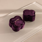 A gold ring with purple stones sits in the Purple Flower Velvet Ring Box, a plush purple velvet box shaped like a blossom, displayed on a glass surface.