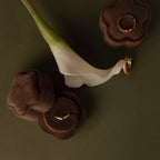 A Brown Flower Velvet Ring Box holds gold rings, elegantly displayed next to a white calla lily on a green surface.