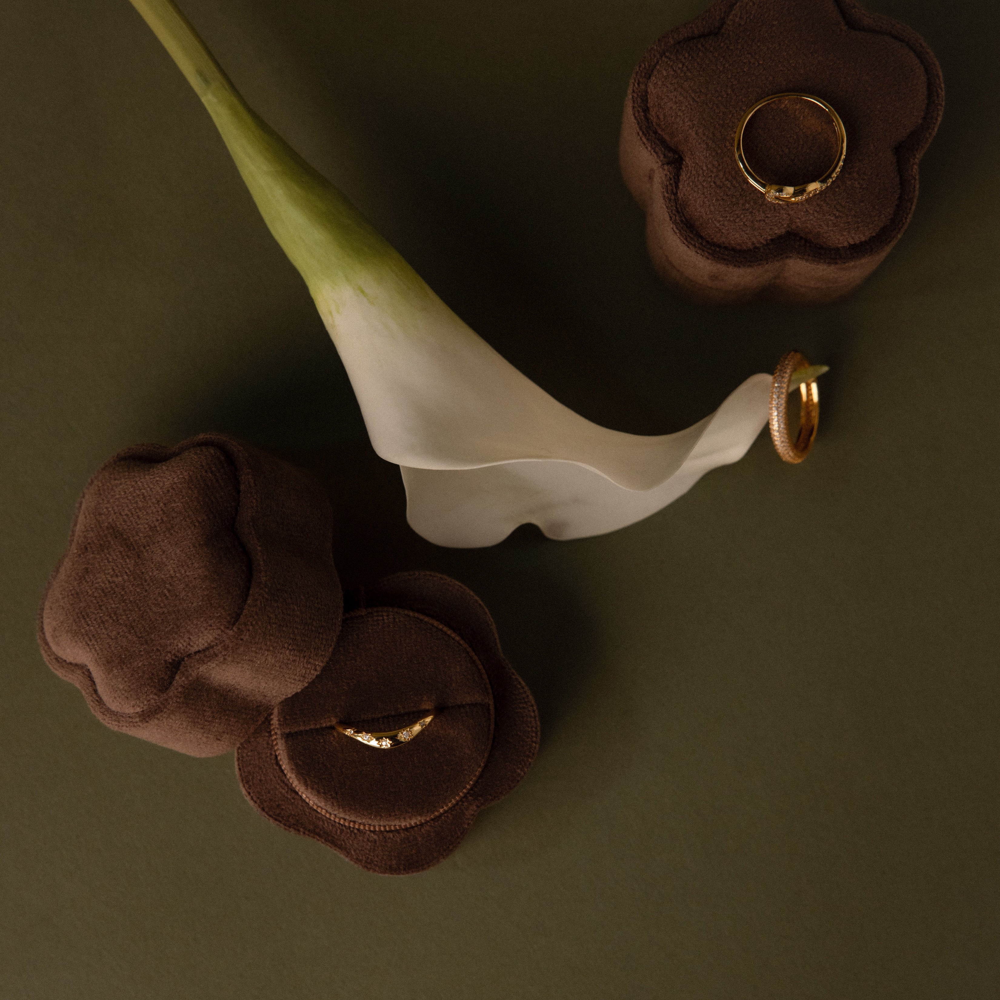 A Brown Flower Velvet Ring Box holds gold rings, elegantly displayed next to a white calla lily on a green surface.