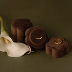 Three Flower Velvet Ring Boxes in brown, each with a gold ring, are displayed beside a white calla lily on an olive green surface—an ideal proposal ring box for your special occasion.