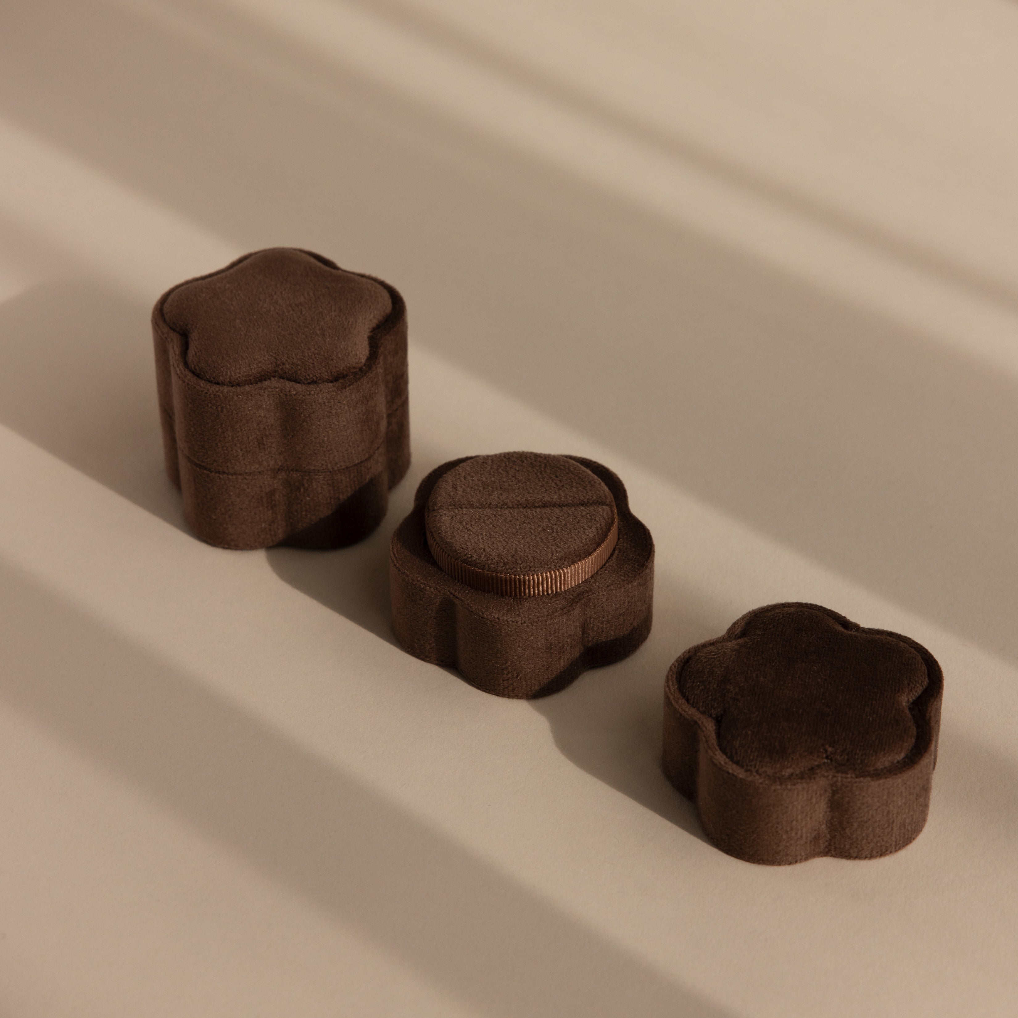 Three Brown Flower Velvet Ring Boxes, featuring a floral design and velvet finish, are arranged on a beige surface with soft shadows.